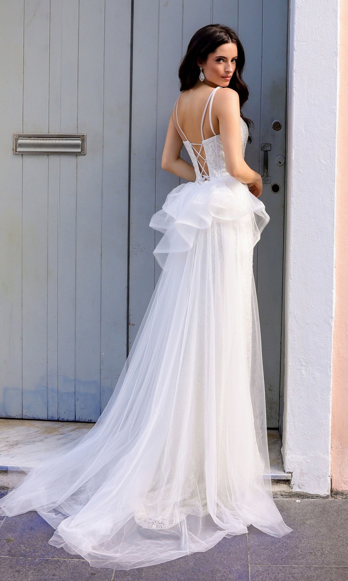 A woman in the Glitter-Print Long Prom Dress with Overskirt Y1475, featuring a backless design and tulle train, stands before a gray door and looks over her shoulder, evoking the elegance of a shimmering evening gown.